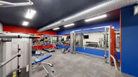 Located at 1960 PA-611 in Swiftwater, Pennsylvania, this dedicated exercise room delivers a fully outfitted fitness environment within a compact, single-floor layout. The space is designed with a bold, energetic aesthetic — vivid red and blue accent walls contrast sharply against crisp white lower walls and a dark ceiling, creating a motivating atmosphere for physical activity.

Overhead, exposed industrial-style ventilation ductwork runs the length of the ceiling alongside recessed LED panel lighting, providing bright, even illumination throughout the workout area. The flooring consists of durable, multi-toned carpet tiles with subtle red accent lines, offering both cushioning and visual interest.

The cardio zone features multiple treadmills and an elliptical trainer, each equipped with individual monitor screens. Full-length mirrors line one wall, visually expanding the space and allowing users to monitor their form. Strength training is well-represented with a Cybex cable machine, a dumbbell rack with a full range of free weights, adjustable benches, and a spin bike.

A wall-mounted water fountain and waste receptacle are conveniently positioned near the entry, which features glass-paneled sliding doors. The thoughtful layout maximizes every square foot, making this exercise room a practical and well-appointed fitness facility.