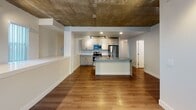 This single-floor space blends industrial character with contemporary finishes to create a stylish and functional living environment. The open-concept layout connects the living area and kitchen seamlessly, anchored by rich hardwood-style flooring that runs throughout the unit. A striking exposed concrete ceiling serves as the defining architectural feature of the main living and kitchen area, lending a raw, loft-like aesthetic that contrasts beautifully with the clean white walls and modern fixtures below. The kitchen is well-appointed with light wood cabinetry, a cool-toned countertop, a tile backsplash, and a full suite of stainless steel appliances including a range, over-the-range microwave, and refrigerator. A central kitchen island with an integrated sink provides additional prep space and doubles as a casual gathering point. Track lighting highlights the kitchen workspace effectively. The bathroom features a large vanity with ample storage, a wide framed mirror, and a distinctive exposed concrete cylindrical column that echoes the industrial theme found elsewhere in the unit. A bedroom offers a ceiling fan, a window providing natural light, and a clean, understated design. The overall ambiance is modern and urban, with thoughtful details that balance industrial elements with comfortable, livable finishes.