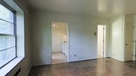 This single-floor space presents a straightforward and functional layout designed for practical living. The main living area features dark hardwood-style flooring that anchors the room, while neutral gray walls create a clean, understated backdrop. The space is open and uncluttered, offering a blank canvas with room to personalize.

An open doorway leads directly into the kitchen, which is illuminated by a pendant light fixture that adds a modest focal point overhead. The kitchen is equipped with a range and what appears to be a washer and dryer unit, making it a compact yet efficient space. Light-toned flooring in the kitchen contrasts with the darker flooring in the main living area, helping to visually distinguish the two zones.

To the right of the main room, a second doorway opens into an additional area, likely a hallway or secondary room, featuring lighter flooring and ample brightness. An electrical panel is mounted on the wall nearby, a practical detail that speaks to the utilitarian nature of the space.

Overall, the space maintains a simple, no-frills character with clean walls, functional fixtures, and a logical flow between rooms.