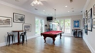 This single-floor game room is a thoughtfully designed recreational space that balances functionality with an inviting atmosphere. The room centers around a handsome billiards table with a bold red felt surface, illuminated by an elegant pendant light overhead. Warm hardwood flooring extends throughout, complemented by neutral gray walls adorned with framed sports memorabilia, pennants, and vintage photographs that lend the space a spirited, collegiate character. Crown molding along the ceiling adds a refined architectural touch, while a ceiling fan with integrated lighting ensures year-round comfort. Multiple large windows line the walls, flooding the room with natural light and offering views of the surrounding greenery and landscaping. French-style doors provide direct access to an outdoor area, further enhancing the open, airy feel. High-top bar tables with industrial-style metal stools are positioned throughout, creating casual seating for spectators and socializing. For added convenience, beverage and snack vending machines are tucked neatly into one corner. Recessed lighting supplements the natural light, ensuring the space remains well-lit at all hours. The overall ambiance is lively yet comfortable, making this game room a versatile hub for entertainment and community engagement.