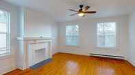 This single-floor unit showcases a blend of classic architectural character and practical design. The living room serves as a focal point, featuring a white-painted decorative fireplace with a traditional mantel, warm honey-toned hardwood floors, and a ceiling fan with integrated lighting. Two double-hung windows frame views of surrounding greenery, filling the space with natural light. Elegant arched doorways connect the living area to additional rooms, adding a distinctive architectural touch.

A separate dining area continues the hardwood flooring and is illuminated by a multi-arm chandelier, with two windows providing ample daylight and neighborhood views. Baseboard heating is present throughout. The bedrooms vary in finish — some feature the same hardwood floors, while one is carpeted — each with multiple windows and ceiling fans or dedicated lighting.

The bathroom is notably detailed, with classic black-and-white patterned mosaic tile surrounding a full soaking tub, marble-style hexagonal floor tile, and a window providing natural ventilation. The kitchen offers white cabinetry, dark countertops, a stainless steel sink with tile backsplash, and hexagonal flooring, completing this characterful space.
