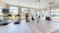 Located at 7600 S Mason Rd in Richmond, TX, this fitness center offers a bright and well-appointed exercise room designed to support a full range of workout routines. The space is finished with light wood-look flooring throughout, contributing to a clean and modern aesthetic that pairs well with the warm neutral wall tones.

Large, floor-to-ceiling black-framed windows line the walls, flooding the interior with abundant natural light and providing open views of the surrounding outdoor landscape. A ceiling fan ensures comfortable airflow during workouts. Recessed lighting supplements natural light for consistent illumination throughout the day.

The exercise room is equipped with a comprehensive selection of fitness equipment, including a treadmill, elliptical trainer, stationary cycle, rowing machine, and a multi-function cable and weight machine. A dedicated free weights area features a full dumbbell rack paired with an adjustable workout bench, all set on protective rubber flooring mats. A wall-mounted television adds to the functionality of the space.

Full-length mirrors along one wall enhance the sense of openness and allow users to monitor their form during strength training. Dark-framed glass panel doors provide both style and accessibility. The overall design balances practicality with a polished, contemporary finish, making this fitness center a functional and inviting space for daily exercise.
