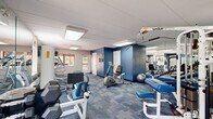 Located at 96 Mary Fran Dr in West Chester, PA, this fitness room provides a dedicated, single-floor workout facility designed to accommodate a comprehensive range of exercise needs. The space features a neutral color palette accented by bold blue walls, with blue-toned modular carpet tiles covering the floor throughout.

The room is well-lit by recessed panel lighting and skylight-style ceiling fixtures, creating a bright and energizing atmosphere. Large windows along one wall allow natural light to filter in and provide views of the surrounding greenery outdoors. Full-length wall mirrors span multiple sections of the room, visually expanding the space and allowing users to monitor their form during workouts.

The cardio section is equipped with multiple treadmills, Precor elliptical machines, and a stair climber, all positioned near the windowed wall to take advantage of the natural light and outdoor views. A wall-mounted television is also present for entertainment during cardio sessions.

The strength training area features a TuffStuff cable multi-station machine, adjustable benches, a leg extension/curl machine, a stationary bike, and a rack of dumbbells in varying weights. A stability ball and resistance accessories round out the available equipment. A wall-mounted HVAC unit ensures climate control throughout the space, keeping the environment comfortable year-round.