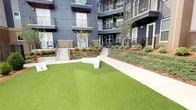 This dynamic communal amenity space combines an energetic indoor lounge with a well-designed outdoor courtyard, offering a range of recreational and social opportunities. The interior is defined by its bold aesthetic: a vivid teal-painted exposed ceiling with industrial ductwork and track lighting sets a lively tone throughout. Wide-plank tile flooring in a neutral wood-look finish grounds the space, while bold artwork and decorative accents add personality to the walls.

The lounge area is thoughtfully appointed with multiple zones for entertainment. A pool table anchors one section, complemented by a sleek wet bar featuring dark countertops, a stainless steel mini-refrigerator, and a teal mosaic tile accent wall with a wall-mounted flat-screen television. A separate media lounge area includes comfortable seating, a large flat-screen display, and a bar-height counter with seating. Large glass doors and windows allow natural light to flow in and provide views of adjacent spaces featuring artistic murals.

The outdoor courtyard is enclosed by a multi-story building with a contemporary dark facade, metal railings, and balconies. Lush artificial turf covers a generous open lawn area, bordered by landscaped beds with shrubs, mulch, and young trees. Concrete pathways and modern angular white benches complete the inviting outdoor setting.