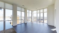 This modern single-floor space makes a striking impression with its expansive floor-to-ceiling windows that wrap around the corner of the main living area, flooding the interior with abundant natural light and offering sweeping panoramic views of the surrounding cityscape. The open-plan layout creates a generous sense of space and flow, ideal for both everyday living and entertaining.

The living area is anchored by rich, dark hardwood flooring that contrasts elegantly with the crisp white walls and bright window frames, lending the space a clean, contemporary aesthetic. The oversized windows are fitted with roller shades, allowing for flexible light control while maintaining the connection to the outdoors.

The elevated position of the unit provides commanding views of the city below, with rooftops, treetops, and urban streets visible in every direction — a standout feature that enhances the overall ambiance of the space. The smooth, neutral-toned ceiling with recessed vents contributes to the minimalist design sensibility throughout.

Overall, this space exudes a bright, airy, and sophisticated character, combining modern architectural details with practical finishes to create an inviting and visually dynamic environment.
