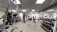 This fitness center is a thoughtfully designed, single-floor exercise facility offering a comprehensive workout environment. The space is brightened by a combination of large recessed panel ceiling lights and natural light streaming in through multiple windows, creating a well-lit and energizing atmosphere. Neutral-toned, textured carpet tiles cover the floor throughout, providing a comfortable and functional surface underfoot.

The facility is outfitted with a diverse selection of cardio equipment, including a treadmill, elliptical trainer, and upright stationary bike, allowing users to engage in a variety of aerobic workouts. A multi-function cable machine serves as the centerpiece for strength and resistance training, complemented by a dedicated weight bench and a well-stocked dumbbell rack offering a wide range of free weights. Exercise balls are also available for core and flexibility training.

A full-length mirror spans one wall, accented by a ballet-style handrail, which not only adds a sense of openness and depth to the room but also supports stretching and form-checking during workouts. Informational fitness charts are posted near the equipment for user reference. The overall ambiance is clean, functional, and inviting, making this fitness center a practical and accessible space for a complete workout routine.