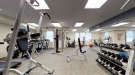 This fitness center is a thoughtfully designed, single-floor exercise facility offering a comprehensive workout environment. The space is brightened by a combination of large recessed panel ceiling lights and natural light streaming in through multiple windows, creating a well-lit and energizing atmosphere. Neutral-toned, textured carpet tiles cover the floor throughout, providing a comfortable and functional surface underfoot.

The facility is outfitted with a diverse selection of cardio equipment, including a treadmill, elliptical trainer, and upright stationary bike, allowing users to engage in a variety of aerobic workouts. A multi-function cable machine serves as the centerpiece for strength and resistance training, complemented by a dedicated weight bench and a well-stocked dumbbell rack offering a wide range of free weights. Exercise balls are also available for core and flexibility training.

A full-length mirror spans one wall, accented by a ballet-style handrail, which not only adds a sense of openness and depth to the room but also supports stretching and form-checking during workouts. Informational fitness charts are posted near the equipment for user reference. The overall ambiance is clean, functional, and inviting, making this fitness center a practical and accessible space for a complete workout routine.