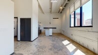 This single-floor space showcases a compelling industrial-modern aesthetic, characterized by dramatically high ceilings with exposed structural beams and a ribbed ceiling panel system. The open layout is anchored by polished concrete floors that carry throughout, lending a raw yet refined character to the interior. Whitewashed brick walls add textural depth while maintaining a clean, bright atmosphere.

The kitchen is a standout feature, equipped with stainless steel appliances including a French door refrigerator and electric range with a stainless hood vent. A central island with a white countertop and integrated sink serves as both a functional workspace and a visual focal point. Open shelving with black iron hardware and wall-mounted swing-arm sconces complete the utilitarian-chic look.

The main living area is expansive and flexible, flooded with natural light from large black-framed windows fitted with roll-down shades. A ceiling fan with track lighting provides adjustable illumination. Louvered utility closet doors line one wall, providing practical storage without interrupting the open flow.

The bathroom features a grey vanity with a stone-topped sink, black fixtures, a framed mirror with a bar light, and polished concrete flooring, maintaining the cohesive industrial palette throughout.