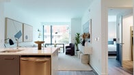 This contemporary single-floor residence showcases a thoughtfully designed open-concept layout with clean lines and a bright, airy aesthetic throughout. Light wood-tone flooring flows seamlessly across the living spaces, complemented by crisp white walls that amplify the abundant natural light pouring in through expansive floor-to-ceiling windows.

The kitchen is outfitted with a full suite of stainless steel appliances, including a gas range, over-the-range microwave, side-by-side refrigerator, and dishwasher. Two-tone cabinetry pairs white upper cabinets with taupe lower drawers, all set against a clean white quartz countertop and backsplash. The kitchen opens directly into the living and dining area, creating an ideal space for entertaining.

The living area benefits from large picture windows that frame urban views and flood the room with daylight. A dedicated seating nook provides a comfortable retreat adjacent to the dining space.

Both bedrooms are generously sized, each featuring oversized windows with city views and distinctive two-tone wall detailing with a deep charcoal lower half. The bathroom is finished with large-format marble-look tile flooring, a sleek white floating vanity, and a wide mirrored medicine cabinet, delivering a polished, spa-like feel.
