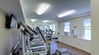 This single-floor fitness center is designed to support a comprehensive cardio workout experience. The space is outfitted with a range of exercise equipment, including stair climbers, stationary bikes, and additional cardio machines, all arranged to maximize usability and flow throughout the room. A full-length mirrored wall runs along one side, visually expanding the space and allowing users to monitor their form during exercise. The flooring is a warm-toned hardwood-style surface, offering both durability and a clean aesthetic. Bright flush-mounted ceiling light panels provide consistent, even illumination throughout the space, supplementing the natural light that enters through multiple windows along the exterior wall. The windows also contribute to ventilation, alongside a wall-mounted air conditioning unit. The walls are finished in a neutral off-white tone, contributing to a clean, clinical, and focused atmosphere. Overall, the fitness center offers a functional and well-maintained environment suited for regular cardiovascular training, with a layout that balances equipment density with comfortable circulation space.