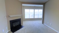 This single-floor space offers a welcoming and functional layout designed for comfortable everyday living. The living area serves as the centerpiece of the space, featuring a sleek, black-framed fireplace with a clean white mantel that provides a classic focal point for the room. Soft, neutral-toned carpeting extends throughout, adding warmth and comfort underfoot. A large, multi-panel window spans much of the exterior wall, flooding the interior with abundant natural light and offering open views of the surrounding outdoor scenery. The walls are finished in a warm, neutral beige tone that complements the overall palette and creates a calm, inviting atmosphere. Crisp white baseboards and trim add a clean, polished contrast throughout the space. The open layout provides a generous sense of space, allowing for flexible furniture arrangements to suit a variety of needs. The combination of natural light, a traditional fireplace, and neutral finishes gives this space a timeless and livable character that is both practical and visually appealing.