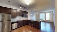 This single-floor space showcases a thoughtfully designed open-concept layout where the kitchen flows seamlessly into the living area, creating a cohesive and functional environment. The kitchen is outfitted with rich, dark espresso cabinetry offering generous storage, complemented by dark granite countertops that extend to a peninsula island — ideal for casual dining or meal prep. Stainless steel appliances, including a built-in microwave, gas range, and dishwasher, provide a sleek, modern aesthetic. A stainless steel undermount sink is positioned beneath the countertop along the main wall, accented by a classic subway tile backsplash. Lighting is a standout feature, with a combination of adjustable track lighting over the kitchen workspace and elegant pendant lights suspended above the peninsula, adding both task and ambient illumination. Warm hardwood flooring runs throughout, grounding the space with a natural, inviting tone. A large window in the adjoining living area allows natural light to filter in, brightening the open layout and enhancing the overall sense of spaciousness.