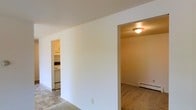 This single-floor residential unit offers a straightforward and functional layout designed for comfortable everyday living. The main living area features tile flooring and is framed by clean white walls that lend a bright, neutral aesthetic throughout the space. From the living area, two distinct openings provide seamless access to the adjoining rooms. To one side, the kitchen is visible through a doorway, showcasing white upper cabinetry, white appliances, and tile flooring that carries a practical, easy-to-maintain character. The kitchen appears well-appointed for daily use, with ample counter space and a cohesive, clean finish. On the opposite side, a carpeted room — suitable as a bedroom or additional living space — is accessible through a second doorway. This room is equipped with a baseboard heater, indicating an efficient heating system, and benefits from a flush-mount ceiling light fixture. The consistent use of white walls throughout the unit creates a cohesive, airy feel that maximizes the sense of space. The overall layout is efficient and well-connected, making it easy to navigate between the living, sleeping, and cooking areas.