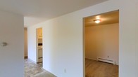 This single-floor residential unit offers a straightforward and functional layout designed for comfortable everyday living. The main living area features tile flooring and is framed by clean white walls that lend a bright, neutral aesthetic throughout the space. From the living area, two distinct openings provide seamless access to the adjoining rooms. To one side, the kitchen is visible through a doorway, showcasing white upper cabinetry, white appliances, and tile flooring that carries a practical, easy-to-maintain character. The kitchen appears well-appointed for daily use, with ample counter space and a cohesive, clean finish. On the opposite side, a carpeted room — suitable as a bedroom or additional living space — is accessible through a second doorway. This room is equipped with a baseboard heater, indicating an efficient heating system, and benefits from a flush-mount ceiling light fixture. The consistent use of white walls throughout the unit creates a cohesive, airy feel that maximizes the sense of space. The overall layout is efficient and well-connected, making it easy to navigate between the living, sleeping, and cooking areas.