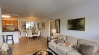 This single-floor residence offers a welcoming and well-designed layout that prioritizes open, connected living. The living area is anchored by a comfortable neutral-toned sofa accented with decorative pillows, complemented by warm hardwood flooring that flows throughout the space. Framed botanical artwork adorns the walls, adding a natural, organic aesthetic to the clean, light-colored interior.

The living space transitions seamlessly into the dining area, which is furnished with a glass-top dining table surrounded by cream upholstered chairs, creating an elegant yet approachable setting for meals and gatherings. A pendant light fixture overhead adds a soft, ambient glow to the dining space, enhancing its inviting atmosphere.

A hallway visible from the main living area leads further into the home, hinting at additional private spaces beyond. The open-plan design allows natural light to travel freely across the rooms, keeping the overall ambiance bright and airy. The consistent use of neutral tones — soft whites, warm beiges, and natural wood — lends a cohesive and serene quality throughout. This thoughtfully styled single-floor space balances functionality with comfort, making it an ideal environment for both everyday living and entertaining.