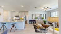 This two-story home is thoughtfully designed to maximize functionality and comfort across both floors. The main level centers around a bright, open-concept living, dining, and kitchen area finished with light hardwood-style flooring. The kitchen is a standout feature, equipped with white shaker-style cabinetry, stainless steel appliances, and a granite-topped island with bar seating — ideal for both everyday meals and entertaining. The adjoining living area is bathed in natural light from multiple windows and is complemented by a ceiling fan overhead. A dedicated workspace on the main floor features large windows that allow ample natural light, making it a practical and inviting area for productivity. Upstairs, the generously sized primary bedroom offers plush carpeting, a full closet, and access to a well-appointed en-suite bathroom featuring a wide dual-sink vanity with quartz countertops and marble-style tile flooring. Two additional bedrooms are also located on the upper level, each with soft carpet and closet space. A versatile bonus area on the upper floor provides extra room that can serve as a lounge or recreation space. Three full bathrooms serve the home with clean, modern finishes throughout. The cohesive grey-and-white palette across the home lends a contemporary, polished ambiance.