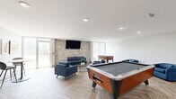 Located at 1722 N Walnut St in Bloomington, Indiana, this single-floor community lounge offers a well-appointed and versatile gathering space designed for both relaxation and recreation. The room is anchored by a striking stacked-stone accent wall, which provides a warm, textural contrast against the crisp white walls and smooth ceiling with recessed lighting. A large flat-screen television is mounted on the stone wall, creating a natural focal point for the lounge seating area, which features a blue sofa, loveseat, and multiple accent chairs arranged around a sleek round coffee table. The herringbone-patterned wood-look tile flooring runs throughout, adding a sophisticated design element that ties the space together. Floor-to-ceiling windows and glass doors allow generous amounts of natural light to flood the interior while offering views to the outdoors. A foosball table and an air hockey table provide dedicated recreation zones, making the space ideal for social engagement. Open black metal shelving units offer display and storage functionality, while a small bistro-style table with modern chairs provides an intimate seating option. The overall ambiance is contemporary and inviting, blending clean architectural lines with comfortable, casual furnishings suited for a communal environment.