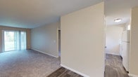 Located at 601 Wenger Rd. in Englewood, Ohio, this single-floor unit offers a practical and comfortable layout throughout. The living area is generously sized, featuring plush carpeting and neutral-toned walls that create a bright, welcoming atmosphere. A sliding glass door floods the space with natural light while providing direct access to an outdoor patio area with green surroundings. A standout architectural detail is the full-height painted brick accent wall that adds texture and character to the main living space, which transitions between carpeted and hard-surface flooring zones. The kitchen is efficiently designed with white cabinetry accented by gold-tone hardware, an electric coil range, an overhead ventilation hood, and hard-surface flooring. The bedroom features soft carpeting and warm neutral walls, offering a quiet and comfortable retreat. The full bathroom includes a vanity with a large mirror, a toilet, and a walk-in shower stall with clean white tile surround. Throughout the unit, white baseboards provide a clean finish, and the overall color palette of warm beiges and soft neutrals contributes to a cohesive, move-in-ready aesthetic. This unit combines functional design with essential amenities in a straightforward, well-maintained layout.