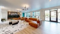 Located at 3219 Manor Rd in Austin, TX, this single-floor community space offers a thoughtfully designed open-concept layout that seamlessly blends a fully equipped kitchen with an expansive living and lounge area. The light wood flooring flows throughout, creating a warm and cohesive aesthetic that ties together each distinct zone.

The kitchen is a standout feature, boasting grey shaker-style cabinetry with gold hardware, a large central island with seating for multiple guests, pendant lighting with gold accents, and floor-to-ceiling marble-look tile. Open wood shelving lines the walls, displaying greenery and decorative accessories. Stainless steel appliances, including a built-in oven and full-size refrigerator, complete this functional yet elegant space.

The living area is generously proportioned, anchored by a bold cognac leather sectional sofa atop a graphic area rug. Statement chandeliers with mixed-metal finishes illuminate the space, while large windows invite abundant natural light and views of the surrounding exterior. A dedicated media nook features a large flat-screen TV and sage-green cabinetry.

A game and lounge zone adds further character, highlighted by a dark feature wall adorned with a curated gallery of black-and-white photography, emerald green banquette seating, a billiards table, and globe pendant lighting — all reflecting Austin's vibrant cultural spirit.