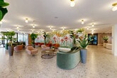 Located in Miami, FL, this generously sized single-floor space makes an immediate impression with its vibrant, resort-inspired aesthetic. The centerpiece of the main lobby is a dramatic floor-to-ceiling tropical botanical mural, bursting with colorful florals and lush foliage that sets a bold, Miami-appropriate tone. Throughout the space, polished terrazzo flooring with multicolored aggregate adds a timeless, elegant foundation that ties each area together seamlessly.

The lounge areas are thoughtfully arranged with curated, modern furnishings — including curved sage-green velvet sofas, cognac leather armchairs, blush pink accent chairs, and brass-legged coffee tables — creating inviting conversation clusters that are both stylish and functional. Tall potted tropical plants are strategically placed throughout, reinforcing the lush, indoor-garden ambiance.

Globe pendant and flush-mount brass light fixtures illuminate the space warmly, while floor-to-ceiling sheer curtains diffuse abundant natural light near the building's entryways. A sculptural, gold-framed multi-panel mirror installation serves as an elegant room divider, adding architectural interest and depth.

A dedicated co-working zone features individual workstations, while a well-stocked convenience store with shelved pantry goods and a refrigerated display provides practical, everyday amenities for occupants. The overall atmosphere is polished, welcoming, and distinctly Miami in character.