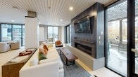 This thoughtfully designed single-floor space showcases a contemporary open-plan layout that seamlessly connects multiple functional zones. Light hardwood flooring runs throughout most of the space, while a distinctive geometric-patterned tile anchors the kitchen area, adding visual contrast and defining the zone.

The kitchen features a long, white waterfall island with a professional-grade faucet, pendant lighting overhead, and a sleek range hood, making it a central and functional focal point. Adjacent to the kitchen, a corridor lined with blue paneled walls and framed artwork leads to additional areas of the space.

A built-in shelving unit with navy blue lower cabinetry and open upper shelves provides generous storage and display opportunities near the entryway, incorporating textured glass panels for added visual interest.

The lounge area is anchored by a linear gas fireplace set within a dark shiplap feature wall, topped by a wall-mounted television. Ample seating and a slatted white ceiling detail add warmth and texture. Floor-to-ceiling windows flood this area with natural light and provide open outdoor views, while a secondary seating nook near the windows offers a quiet retreat within the larger space.