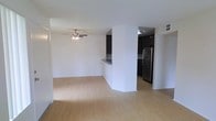 This single-floor space presents an open-concept layout that seamlessly connects the living area and kitchen. The living area is finished with light hardwood-style flooring that extends throughout, creating a bright, cohesive look. A ceiling fan with a light fixture is centrally positioned overhead, providing both ventilation and ambient lighting to the main living space.

The kitchen is partially separated by a half-wall partition, which also serves as a breakfast bar-style counter. Dark cabinetry contrasts sharply with the white walls, lending the kitchen a contemporary aesthetic. A stainless steel refrigerator anchors the kitchen area, complementing the dark cabinet finishes. Recessed lighting illuminates the kitchen, adding to its functional appeal.

The white walls throughout the space contribute to a clean, neutral palette that enhances the sense of openness. The layout allows for flexible furniture arrangement in the living area, while the kitchen's partial enclosure provides definition without sacrificing connectivity. The overall design is modern and practical, suited for everyday living.