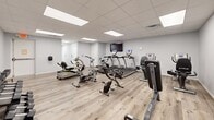 Located at 1922 W 161st St S in Glenpool, Oklahoma, this single-floor amenity space combines an active fitness area with a welcoming lounge, offering a well-rounded environment for both exercise and relaxation.

The exercise room is bright and open, finished with light wood-tone flooring and clean white walls that give it a crisp, motivating atmosphere. Illuminated by recessed panel lighting set into a drop-tile ceiling, the space is outfitted with a comprehensive selection of cardio and strength equipment, including treadmills, stationary bikes, an elliptical, a rowing machine, a weight bench, and a dumbbell rack — providing a thorough workout experience within a single room. A wall-mounted TV keeps users entertained during their sessions.

Adjacent to the fitness area, the living room offers a warm contrast with its deep blue-gray accent walls and stylish sconce lighting that creates an inviting, relaxed ambiance. Comfortable leather recliner chairs, a gray sofa, and a mustard accent chair are arranged around a patterned area rug, centered on a wall-mounted television. The thoughtfully chosen furnishings and soft lighting make this lounge an ideal space for unwinding or socializing. Both rooms share the same durable wood-look flooring, tying the entire space together with a cohesive, modern aesthetic.