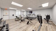 Located at 1922 W 161st St S in Glenpool, Oklahoma, this single-floor amenity space combines an active fitness area with a welcoming lounge, offering a well-rounded environment for both exercise and relaxation.

The exercise room is bright and open, finished with light wood-tone flooring and clean white walls that give it a crisp, motivating atmosphere. Illuminated by recessed panel lighting set into a drop-tile ceiling, the space is outfitted with a comprehensive selection of cardio and strength equipment, including treadmills, stationary bikes, an elliptical, a rowing machine, a weight bench, and a dumbbell rack — providing a thorough workout experience within a single room. A wall-mounted TV keeps users entertained during their sessions.

Adjacent to the fitness area, the living room offers a warm contrast with its deep blue-gray accent walls and stylish sconce lighting that creates an inviting, relaxed ambiance. Comfortable leather recliner chairs, a gray sofa, and a mustard accent chair are arranged around a patterned area rug, centered on a wall-mounted television. The thoughtfully chosen furnishings and soft lighting make this lounge an ideal space for unwinding or socializing. Both rooms share the same durable wood-look flooring, tying the entire space together with a cohesive, modern aesthetic.