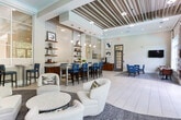 Located at 1193 Collier Rd NW in Atlanta, Georgia, this single-story community clubhouse is thoughtfully designed to blend comfort, functionality, and modern aesthetics. The open-plan layout flows seamlessly across light-toned large-format tile flooring, creating a bright and welcoming atmosphere throughout.

The lounge area serves as a focal point of the space, anchored by a floor-to-ceiling tiled fireplace surround with built-in display niches and a wall-mounted flat-screen television. Plush white swivel chairs and a coffee table invite relaxed conversation in this elegantly appointed sitting area.

A dedicated bar and café zone features a sleek counter lined with bold blue upholstered high-back bar stools, complemented by a textured mosaic tile accent wall with open wooden shelving. Glass partition walls allow light to pass freely between zones while maintaining a sense of definition within the space.

Multiple seating arrangements accommodate both social and individual use, including café-style tables paired with richly toned leather chairs arranged atop area rugs. Large multi-pane windows dressed with patterned drapery flood the interior with natural light and frame views of lush greenery and the outdoor pool area, which is accessible through glass double doors. The overall ambiance is refined yet approachable, making it an ideal communal gathering space.