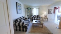 This three-floor residence offers a well-organized layout spread across multiple levels, providing generous separation between living and private spaces. The living room, pictured here, serves as the central gathering area and is furnished with bold black-and-white buffalo check upholstered seating, including a full sofa and loveseat arranged around a wooden coffee table. A neutral-toned area rug anchors the seating arrangement atop wall-to-wall carpeting, adding warmth and texture to the room. A window dressed with sheer curtains allows soft, diffused natural light to fill the space, contributing to an airy and relaxed atmosphere. Decorative touches include a framed abstract artwork on the wall and a metal etagere displaying a potted plant, adding visual interest and a touch of greenery. A table lamp provides warm supplemental lighting for evening use. The overall aesthetic of the living area is casual and comfortable, with a classic sensibility. The multi-floor structure of the home suggests additional rooms and functional spaces across the remaining levels, offering ample room for everyday living.