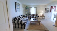 This three-floor residence offers a well-organized layout spread across multiple levels, providing generous separation between living and private spaces. The living room, pictured here, serves as the central gathering area and is furnished with bold black-and-white buffalo check upholstered seating, including a full sofa and loveseat arranged around a wooden coffee table. A neutral-toned area rug anchors the seating arrangement atop wall-to-wall carpeting, adding warmth and texture to the room. A window dressed with sheer curtains allows soft, diffused natural light to fill the space, contributing to an airy and relaxed atmosphere. Decorative touches include a framed abstract artwork on the wall and a metal etagere displaying a potted plant, adding visual interest and a touch of greenery. A table lamp provides warm supplemental lighting for evening use. The overall aesthetic of the living area is casual and comfortable, with a classic sensibility. The multi-floor structure of the home suggests additional rooms and functional spaces across the remaining levels, offering ample room for everyday living.