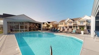 Located in Hilliard, Ohio, this single-level community amenity space centers around a generously sized outdoor swimming pool with crystal-clear blue water, making it a standout feature of the property. The pool area is surrounded by a well-maintained concrete deck furnished with a row of cushioned lounge chairs, providing ample space for sunbathing and relaxation. Safety amenities, including a life ring station, are thoughtfully integrated into the pool surround. Adjacent to the pool, a screened pavilion offers a shaded, enclosed gathering area with views of the pool deck — ideal for outdoor socializing while staying protected from the elements. The overall design of the amenity space reflects a clean, resort-inspired aesthetic with a welcoming, open-air atmosphere. The surrounding landscape is framed by multi-story residential buildings with classic architectural detailing, including neutral siding, pitched rooflines, and balconies that overlook the community. Mature trees and black iron perimeter fencing add structure and a sense of privacy to the space. Under open skies, this outdoor amenity area delivers a bright, airy environment well-suited for community gatherings and warm-weather recreation in the heart of a Hilliard neighborhood.