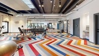 Located at 801 Camino del Rio in Durango, Colorado, this fitness center offers a well-appointed and energetic workout environment designed to motivate and accommodate a wide range of exercise routines. The space is defined immediately by its bold, multicolored chevron-patterned flooring in red, blue, orange, and gray tones, lending the room a dynamic, modern aesthetic that sets it apart from a typical gym setting.

The ceiling features exposed dark-painted structural beams with integrated track lighting, adding an industrial-chic character while ensuring the space is well-lit throughout. A central skylight floods the interior with natural light, enhancing the open and airy atmosphere. Large glass doors and windows along one wall further connect the interior to the outdoors and provide additional natural illumination.

The fitness floor is comprehensively equipped with cardio machines including treadmills, ellipticals, and stationary cycling bikes, as well as a full cable strength machine, a dumbbell rack with adjustable benches, and stability balls for functional training. A mirrored wall section expands the sense of space and supports form-checking during workouts. Lockers and informational signage are also present, adding practical utility. The layout provides ample open floor space for stretching or floor-based exercises, making this fitness center a complete and thoughtfully designed facility.