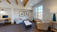 Located at 287 Huger St in Charleston, South Carolina, this thoughtfully designed single-floor amenity space blends Southern charm with modern sophistication. Exposed natural wood beams run the length of the ceiling, creating warmth and architectural character throughout the open layout. The flooring is covered in a tasteful patterned carpet that ties the space together cohesively.

A comfortable lounge area anchors one end of the space, featuring neutral upholstered armchairs and a sofa arranged around a marble-topped coffee table. A large multi-pane window floods this area with natural light and offers views of the surrounding neighborhood. Framed bird artwork adorns the walls, adding a refined, curated aesthetic.

Toward the center of the space, a built-in business workstation provides a functional nook equipped with a desktop computer, printer, and a warm-toned wood work surface accented by wall-mounted task lighting and coastal-themed artwork above.

Prominent steel-framed glass double doors open to reveal a dramatic stairwell overlook, complete with a striking sculptural chandelier and a bold teal built-in shelving unit with wood accents. The iron railing with a wood cap rail frames the open staircase below, adding an industrial-yet-refined touch. Recessed lighting throughout ensures the space remains bright and welcoming at all times.