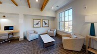 Located at 287 Huger St in Charleston, South Carolina, this thoughtfully designed single-floor amenity space blends Southern charm with modern sophistication. Exposed natural wood beams run the length of the ceiling, creating warmth and architectural character throughout the open layout. The flooring is covered in a tasteful patterned carpet that ties the space together cohesively.

A comfortable lounge area anchors one end of the space, featuring neutral upholstered armchairs and a sofa arranged around a marble-topped coffee table. A large multi-pane window floods this area with natural light and offers views of the surrounding neighborhood. Framed bird artwork adorns the walls, adding a refined, curated aesthetic.

Toward the center of the space, a built-in business workstation provides a functional nook equipped with a desktop computer, printer, and a warm-toned wood work surface accented by wall-mounted task lighting and coastal-themed artwork above.

Prominent steel-framed glass double doors open to reveal a dramatic stairwell overlook, complete with a striking sculptural chandelier and a bold teal built-in shelving unit with wood accents. The iron railing with a wood cap rail frames the open staircase below, adding an industrial-yet-refined touch. Recessed lighting throughout ensures the space remains bright and welcoming at all times.