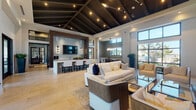 This upscale communal clubhouse is designed to impress, offering a cohesive blend of modern elegance and comfortable functionality across a single, open floor plan. The space is anchored by dramatic vaulted ceilings with dark exposed beams and integrated track lighting, creating a warm yet sophisticated atmosphere throughout.

The main lounge area features generous seating arrangements with contemporary cream-toned sofas, armchairs, and glass-top coffee tables set atop textured area rugs over large-format tile flooring. A striking tiled accent wall serves as the backdrop for a mounted flat-screen television and a sleek bar counter with decorative patterned stools, making it an ideal gathering spot.

A secondary lounge zone showcases a floor-to-ceiling herringbone-patterned stone feature wall framing a linear gas fireplace and another mounted screen, surrounded by plush seating—perfect for relaxed conversation or viewing. Nearby, a game area with a full-length wooden shuffleboard table and additional casual seating adds a lively recreational element, enhanced by large windows that flood the space with natural light and views of the surrounding outdoor landscape.

The bright, airy entryway welcomes visitors with soaring ceilings, expansive glass doors and windows, and clean white walls that set an inviting tone. Throughout the space, thoughtful architectural detailing, layered lighting, and high-end finishes combine to deliver a refined yet approachable environment.