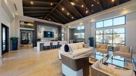 This upscale communal clubhouse is designed to impress, offering a cohesive blend of modern elegance and comfortable functionality across a single, open floor plan. The space is anchored by dramatic vaulted ceilings with dark exposed beams and integrated track lighting, creating a warm yet sophisticated atmosphere throughout.

The main lounge area features generous seating arrangements with contemporary cream-toned sofas, armchairs, and glass-top coffee tables set atop textured area rugs over large-format tile flooring. A striking tiled accent wall serves as the backdrop for a mounted flat-screen television and a sleek bar counter with decorative patterned stools, making it an ideal gathering spot.

A secondary lounge zone showcases a floor-to-ceiling herringbone-patterned stone feature wall framing a linear gas fireplace and another mounted screen, surrounded by plush seating—perfect for relaxed conversation or viewing. Nearby, a game area with a full-length wooden shuffleboard table and additional casual seating adds a lively recreational element, enhanced by large windows that flood the space with natural light and views of the surrounding outdoor landscape.

The bright, airy entryway welcomes visitors with soaring ceilings, expansive glass doors and windows, and clean white walls that set an inviting tone. Throughout the space, thoughtful architectural detailing, layered lighting, and high-end finishes combine to deliver a refined yet approachable environment.