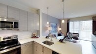 This well-appointed 1-bedroom, 1-bathroom residence is designed with a modern aesthetic and a thoughtful open-concept layout that connects the kitchen and living areas seamlessly. The kitchen is a standout feature, equipped with stainless steel appliances including a refrigerator, electric range, and built-in microwave. Light gray flat-panel cabinetry with bar-pull hardware extends floor to ceiling, offering generous storage, while a classic subway tile backsplash adds texture and visual interest. White quartz countertops provide ample prep space, and a peninsula with a stainless steel sink anchors the space. Elegant pendant lights hang above the peninsula, reinforcing the kitchen's contemporary character. Light wood-tone flooring runs throughout the kitchen and into the living area, enhancing the sense of continuity. The living room is bright and welcoming, bathed in natural light from large sliding glass doors fitted with vertical blinds. The bedroom is a calm retreat, featuring a neutral color palette accented by a deep gray feature wall, natural light from windows with blinds and drapes, and ample space for a full furniture arrangement. Throughout the residence, a cohesive palette of soft grays, whites, and warm neutrals creates a polished and inviting atmosphere.