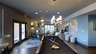 Located at 3001 Market Center Dr in Morrisville, NC, this single-floor community clubhouse offers a sophisticated and inviting atmosphere designed for both relaxation and social engagement. The space is anchored by a full-size billiards table positioned beneath a striking cluster of cylindrical pendant chandeliers, creating a focal point that sets a refined yet playful tone. Recessed lighting and wall-mounted sconces layer warm, ambient illumination throughout the room, complementing the dark hardwood flooring and neutral color palette. Multiple distinct lounge seating areas are arranged thoughtfully across the space, each furnished with upholstered accent chairs and complementary area rugs, evoking a sense of intimate comfort within a larger, open layout. A fireplace adds a welcoming architectural element to one of the seating zones, enhancing the cozy ambiance. Curved architectural details and sweeping walls contribute a sense of dynamic flow to the floor plan. Large windows and glass doors allow ample natural light to filter into the interior, connecting the space visually to the outdoors. Curated artwork adorns the walls, adding a gallery-like quality that elevates the overall aesthetic. This well-appointed clubhouse space is designed to foster community while delivering a polished, upscale experience.