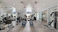 This fitness center is a thoughtfully designed, single-floor facility that offers a spacious and welcoming environment for exercise. The architecture is a standout feature, highlighted by soaring vaulted ceilings with a striking arched window that floods the main workout area with natural light. Ceiling fans are suspended overhead, promoting airflow throughout the space, while recessed lighting ensures consistent illumination at all hours.

The main workout floor is finished with a patterned, chevron-style carpet tile in neutral tones of gray and beige, providing both comfort and durability underfoot. The open layout accommodates a generous array of cardio equipment, including multiple treadmills, elliptical machines, stationary bikes, and stair climbers. Mounted televisions are positioned for easy viewing during cardio sessions.

A dedicated strength training area features selectorized cable machines and weight equipment, partially separated by frosted glass sliding panels that add a modern architectural touch. Full-length mirrors line portions of the walls, enhancing the sense of space and allowing users to monitor their form. A quieter annex room provides additional strength training space with natural views through large windows. The overall ambiance is clean, bright, and professional, creating an inviting atmosphere for a complete fitness experience.