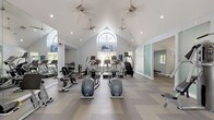 This fitness center is a thoughtfully designed, single-floor facility that offers a spacious and welcoming environment for exercise. The architecture is a standout feature, highlighted by soaring vaulted ceilings with a striking arched window that floods the main workout area with natural light. Ceiling fans are suspended overhead, promoting airflow throughout the space, while recessed lighting ensures consistent illumination at all hours.

The main workout floor is finished with a patterned, chevron-style carpet tile in neutral tones of gray and beige, providing both comfort and durability underfoot. The open layout accommodates a generous array of cardio equipment, including multiple treadmills, elliptical machines, stationary bikes, and stair climbers. Mounted televisions are positioned for easy viewing during cardio sessions.

A dedicated strength training area features selectorized cable machines and weight equipment, partially separated by frosted glass sliding panels that add a modern architectural touch. Full-length mirrors line portions of the walls, enhancing the sense of space and allowing users to monitor their form. A quieter annex room provides additional strength training space with natural views through large windows. The overall ambiance is clean, bright, and professional, creating an inviting atmosphere for a complete fitness experience.