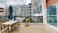 Located at 4359 Sadler Rd in Glen Allen, Virginia, this single-story amenity space is set within a contemporary multi-story residential development and designed to foster an engaging outdoor and indoor community experience. The exterior courtyard serves as the heart of the space, featuring herringbone-patterned brick pavers that define a generous patio area furnished with seating, granite-topped communal tables, and string lights that add ambient character. A well-manicured lawn extends alongside the patio, dotted with Adirondack chairs and bordered by landscaped planters, creating an inviting open-air gathering area. Outdoor televisions and shaded seating clusters are positioned near the lawn, enhancing the space's utility for both casual lounging and social events. An ornamental iron fence lines one edge of the patio, offering views of an adjacent pool area and manicured greenery beyond. The indoor component is accessed through floor-to-ceiling glass garage-style doors that fold open to blur the boundary between interior and exterior. Inside, warm wood-look flooring, blue tile accents, and pendant lighting create a polished yet approachable atmosphere. The surrounding multi-story building facades, clad in a mix of gray siding, warm wood tones, and masonry, frame the courtyard and contribute to a cohesive, modern aesthetic throughout the space.