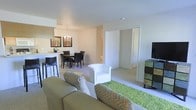 This single-floor residence features an open-concept layout that seamlessly connects the living, dining, and kitchen areas into one cohesive space. The living area is anchored by a plush sofa adorned with patterned throw pillows and paired with a white accent chair, all set atop a vibrant green area rug that adds a pop of color to the neutral palette. A flat-screen television sits on a contemporary media console along one wall, completing the entertainment setup.

The dining area transitions naturally from the living space, featuring a dark wood table surrounded by matching upholstered chairs beneath an elegant chandelier that serves as a focal point. Adjacent to the dining area, a raised breakfast bar with bar seating provides additional casual dining options. Wall art and a small floral arrangement add decorative touches to this zone.

The kitchen is accessible from the dining area and shares the open layout. Neutral walls and light carpeting run throughout, contributing to the bright and airy atmosphere. A hallway visible from the living area leads to additional rooms, suggesting a well-organized and functional floor plan. The overall ambiance is clean, welcoming, and thoughtfully arranged.
