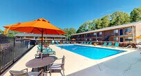 Located at 404 Jones Ferry Rd in Carrboro, North Carolina, this outdoor pool area serves as a central community amenity surrounded by classic brick residential buildings set against a lush, tree-lined backdrop. The expansive swimming pool features clear, blue water and is designed for recreational use, accommodating multiple swimmers comfortably. The generous concrete pool deck wraps around the perimeter and is furnished with numerous teal lounge chairs, providing ample space for sunbathing and relaxation. Vibrant orange patio umbrellas shade seating areas with tables and chairs, adding a lively pop of color to the outdoor environment. Potted flowering plants are placed throughout, enhancing the inviting atmosphere. The pool area is fully enclosed by a wrought iron fence, ensuring privacy and security. Posted safety signage is displayed prominently along the brick building walls, providing clear pool rules and guidelines in both English and Spanish. The surrounding grounds are well-landscaped with neatly trimmed shrubs and mature trees that offer a natural canopy, creating a comfortable and shaded setting. Under sunny North Carolina skies, this pool amenity offers a welcoming and functional outdoor retreat for residents.