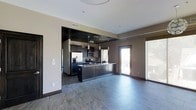 This single-floor residence showcases a thoughtfully designed open-concept layout that seamlessly connects the kitchen and living areas. The kitchen is a standout feature, anchored by a substantial center island with dark cabinetry and granite-style countertops. A dramatic dropped ceiling with dark paneling frames the kitchen zone, creating a distinct yet integrated atmosphere. Stainless steel appliances, including a refrigerator and built-in microwave, complement the space alongside a mosaic tile backsplash that adds textural detail. Pendant lighting hangs above the island, providing both task illumination and visual appeal. The floors throughout are covered in large-format slate-style stone tiles, lending a grounded, upscale character to the interior. Neutral warm-toned walls provide a versatile backdrop throughout the space. A sliding glass door leads directly to an outdoor balcony, allowing natural light to flow into the living area and offering a connection to the exterior. The overall ambiance is modern and polished, with dark finishes contrasting against lighter walls and ceilings to create a sophisticated, contemporary aesthetic suited to everyday living.
