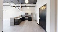 This thoughtfully designed one-bedroom, one-bathroom residence at 480 Myrtle Street in New Britain, CT, blends industrial character with modern finishes throughout its well-proportioned floor plan. The open living and kitchen area immediately impresses with soaring ceilings, exposed ceiling beams and ductwork, and globe pendant lighting that reinforces the loft-inspired aesthetic. Light-toned luxury vinyl flooring runs through the main living spaces, complementing the crisp white walls and bold black accents. The kitchen is appointed with two-tone cabinetry — white upper cabinets paired with sleek black lower cabinets — stainless steel appliances including a gas range, microwave, and refrigerator, and a gold-finish faucet at the sink, adding a refined touch. The spacious bedroom features plush carpeting, a large multi-pane window that floods the room with natural light, and open views of the surrounding landscape. A dedicated closet provides convenient storage. The full bathroom offers a clean, all-white design with a shower/tub combination, tile flooring, a white vanity, and a round black-framed mirror. The unit's bold black entry door with gold hardware makes a striking first impression, tying together the space's cohesive modern-industrial design language.