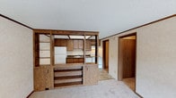 This single-floor home is thoughtfully laid out to maximize functionality and comfort. The living area features neutral carpet and flows openly toward the kitchen, which is separated by a distinctive built-in oak wood shelving and cabinetry unit that doubles as a room divider and display space. This unique architectural element adds character while maintaining an open, connected feel between the two areas.

The kitchen is well-equipped with dark laminate countertops, warm oak cabinetry both above and below the counter, a single-basin stainless steel sink, a white electric coil-top range, a dishwasher, and a refrigerator. Tile flooring grounds the kitchen space and complements the wood tones throughout.

The bedrooms are carpeted and finished with neutral walls, crown molding, and dark wood baseboards. One bedroom features a ceiling fan with light fixture and a window that allows natural light to filter in. Closet space is accessible via wood-paneled doors.

The bathroom is efficiently designed, featuring an oak vanity with a dark laminate countertop, a medicine cabinet mirror, a toilet, and a full tub-shower combo with a clean white surround. Tile flooring ties the room together neatly.

A dedicated laundry room houses a full-size washer and dryer alongside a water heater and electrical panel, providing practical utility within the home. Textured ceilings and consistent trim work appear throughout, giving the space a cohesive, understated aesthetic.