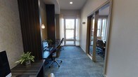 This contemporary business center is thoughtfully designed to support a productive and professional work environment. The main workspace features a built-in dark wood desk running along one wall, equipped with multiple computer workstations and ergonomic rolling chairs, making it ideal for focused individual work or collaborative tasks. Patterned blue-grey carpeting adds a polished, corporate feel underfoot throughout the space.

Natural light flows generously through a large window at the end of the room, framed by sheer white curtains that soften the brightness while maintaining an open, airy atmosphere. Warm pendant lighting and recessed ceiling fixtures complement the natural light, ensuring well-balanced illumination at all hours.

A private office or conference room is accessible through a glass-paneled sliding door along the right wall, offering a more secluded setting for meetings or confidential work. This enclosed space is similarly furnished with a desk and seating, maintaining visual continuity with the main area through its glass partition.

Dark wood wall accents and neutral-toned walls create a refined, sophisticated aesthetic throughout. The overall design strikes a balance between functionality and modern elegance, providing a comfortable and efficient setting for business and professional tasks.