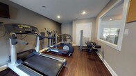 Located at 1660 Old Trolley Rd in Summerville, South Carolina, this single-floor fitness center is a well-equipped facility designed to support a range of workout routines. The space features rich, dark hardwood flooring throughout, complemented by neutral gray walls and recessed lighting that creates a clean, focused environment for exercise.

The cardio area is prominently equipped with multiple Life Fitness treadmills and elliptical trainers, arranged to maximize use of the floor space. Wall-mounted flat-screen televisions are positioned for easy viewing during cardio sessions. A rowing machine provides additional low-impact cardio options, while a multi-function strength training cable machine rounds out the strength equipment offerings.

Natural light filters into the space through windows and a glass-paneled entry door, offering views of the surrounding outdoor area. The entry zone includes a sanitizing station and posted fitness center rules, promoting a clean and organized environment. Decorative yellow abstract artwork adorns one wall near the entrance, adding a subtle pop of color to the otherwise understated interior palette.

The layout efficiently accommodates both cardio and strength training within a compact, single-level footprint, making this fitness center a practical and well-appointed amenity for residents and users of the Summerville location.