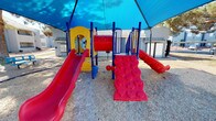 This outdoor playground is a well-equipped recreational space designed to encourage active play for children. The centerpiece is a bold, multi-level play structure finished in bright primary colors — red, yellow, and blue — making it visually engaging and easy for children to navigate. The structure includes multiple red slides of varying sizes, a textured red climbing wall with molded handholds, and a set of stairs with sturdy blue railings for safe access to the elevated platform. Interactive elements such as colorful activity panels and a mirror are integrated into the structure to stimulate imaginative and sensory play. The entire play area is covered by a large, tensioned blue shade canopy supported by tall poles, providing meaningful protection from direct sunlight and making the space comfortable for extended outdoor play. The ground surface beneath the structure is covered with wood chip or gravel material, offering a softer, impact-absorbing layer for safety. The surrounding environment features mature trees and low-maintenance desert landscaping, with residential buildings visible in the background, suggesting this playground is part of a larger community or residential complex. The overall design prioritizes both fun and safety in a well-maintained, accessible outdoor setting.